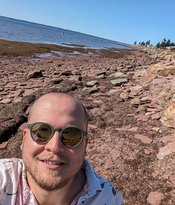 Selfie of Matthew Ludwig, wearing sunglasses, on a rocky shoreline. Ocean visible in the distance.