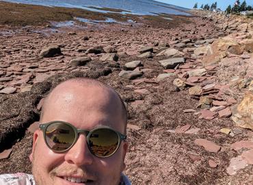 Selfie of Matthew Ludwig, wearing sunglasses, on a rocky shoreline. Ocean visible in the distance.